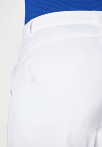 Calças brancas com uma textura suave, apresentando um bolso traseiro e um discreto detalhe de logo bordado. Acompanhadas por um top azul por cima.