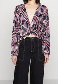 Blusa de manga larga con un escote en V profundo, con nudo en el frente, estampada en púrpura, negro y azul, hecha de una tela ligera; combinada con pantalones de pierna ancha negros.