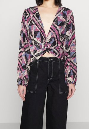 Blusa de manga larga con un escote en V profundo, con nudo en el frente, estampada en púrpura, negro y azul, hecha de una tela ligera; combinada con pantalones de pierna ancha negros.