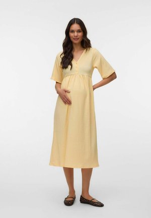 Kobieta w ciąży w żółtej sukience midi z krótkimi rękawami, stoi z jedną ręką na brzuchu, a drugą na biodrze, nosi czarne baleriny.