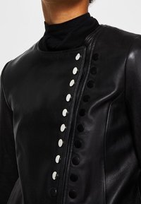 Musta nahkatakki, jossa on korkea kaulus ja hopeiset pyöreät napit etuosassa. Sileä pinta, istuva malli ja minimaaliset saumat.