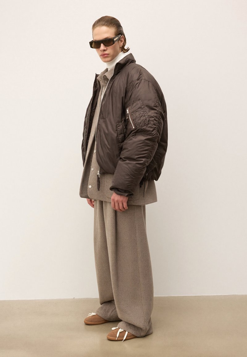 Jachetă bomber căptușită de culoare maro închis, cu un buzunar cu fermoar, purtată peste o cămașă gri deschis. Pantaloni bej largi și sandale maro cu talpă plată completează ținuta.