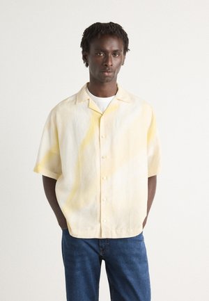 Short-sleeve button-up shirt in a light yellow hue with subtle abstract patterns, made from a soft fabric. Paired with dark blue jeans.
