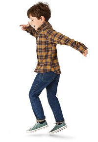 Karierte gelb-braune Langarmbluse, blaue Jeans und grüne Turnschuhe. Das Kind ist in Bewegung, die Arme ausgestreckt.