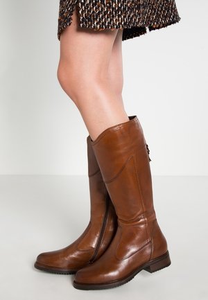 Jambes portant des bottes en cuir marron montant jusqu'aux genoux et une jupe à motif tweed sur un fond clair uni.
