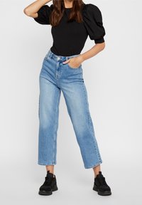 Černý top s balónovými rukávy, zkombinovaný s vysokými světle modrými širokými džíny. Model má na sobě černé tenisky. Hladký materiál a volný střih.