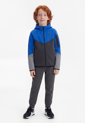 Gyerek vörös hajjal, kék, fekete és szürke Nike kapucnis pulóvert, sötétszürke nadrágot és fekete Nike tornacipőt visel, előre nézve áll.