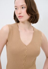 Beige ärmelloses Oberteil mit gerippter Textur, V-Ausschnitt und dekorativen Knopfdetails entlang der Vorderseite. Glattes Material, taillierter Schnitt.