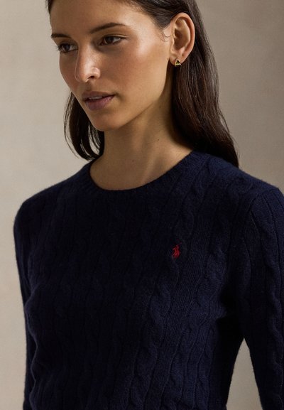 Femme aux cheveux foncés portant des boucles d'oreilles créoles dorées et un pull bleu marine en maille torsadée avec un petit logo rouge brodé sur la poitrine.