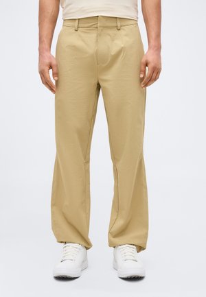 Άνδρας που φοράει μπεζ παντελόνι κανονικής γραμμής και λευκά αθλητικά παπούτσια, στέκεται μπροστά σε απλό φωτεινό φόντο.