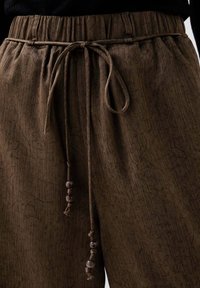 Bruine plooibroek met een elastische tailleband en houten kralenaccenten. Getextureerde stof met een subtiele designdetail.