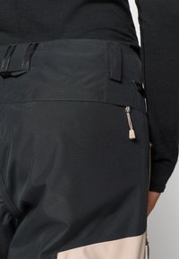 Pantaloni din material negru cu un panou contrastant bej. Prezintă o textură netedă, un buzunar utilitar cu fermoar și bretele ajustabile pe talie.