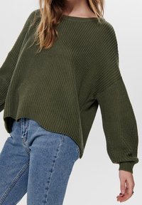 Pull surdimensionné en maille côtelée de couleur vert olive. Il présente des manches larges, un col bateau et un ourlet arrière légèrement allongé. Porté avec un jean bleu.