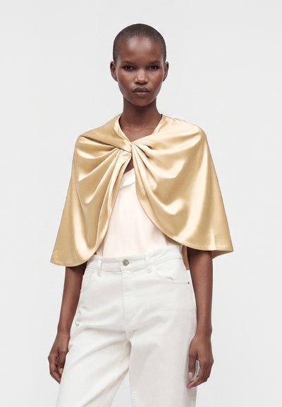 Femme aux cheveux courts portant une cape en satin doré brillant par-dessus un haut blanc et un pantalon blanc sur un fond clair uni.
