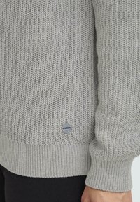 Pull gris tricoté avec un motif texturé, des poignets et un ourlet côtelés, arborant un petit logo circulaire sur le côté gauche.