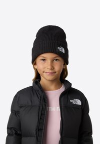 Μαύρο ριμπ πλεκτό σκουφί με ετικέτα λογότυπου, φορεμένο με μαύρο φουσκωτό μπουφάν και ανοιχτό ροζ πουκάμισο με λευκό κείμενο.