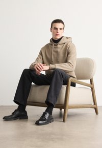 Felpa beige con cappuccio e lacci, dolcevita nero, jeans neri e scarpe stringate nere. Seduto su una sedia di legno curvata con un cuscino testurizzato.