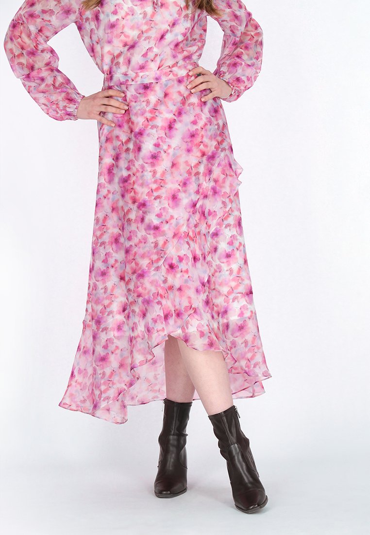Person, die ein pinkes und lila blumiges High-Low-Kleid mit langen Ärmeln und schwarzen Stiefeletten trägt, die Hände auf den Hüften.