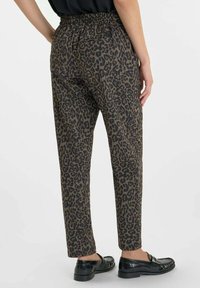 Persoon in hoge taille, taps toelopende broek met luipaardprint en zwarte instappers, staand tegen een effen lichte achtergrond.