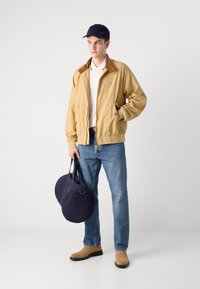 Beige bomberjakke med brun krave, hvid polo skjorte, blå jeans og lysebrune støvler; holder to mørkeblå runde tasker.