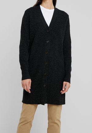 Personne portant un long cardigan noir côtelé boutonné sur un haut blanc et un pantalon beige sur un fond gris uni.
