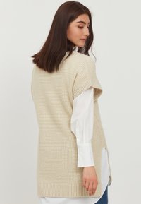Gilet sans manches en beige clair, avec un motif texturé, un ourlet côtelé et des fentes sur les côtés. Superposé sur une chemise blanche à col.