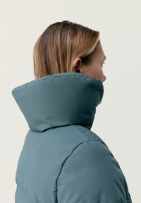 Vadderad jacka i teal med en hög, strukturerad krage. Slät, matt yta med en ren design och subtila sömnadsdetaljer.