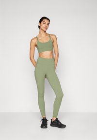 Green sports bra and matching high-waisted leggings made of stretchy fabric. Black athletic shoes. Smooth texture, minimal design.