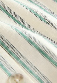 Gewebter Stoff mit cremefarbigem Hintergrund und dünnen Streifen in Türkis und Marineblau. Mit einem klaren Knopfdetail. Strukturierte Oberfläche.