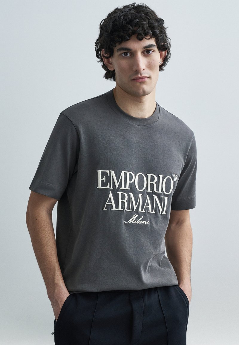Sivé bavlnené tričko s veľkým bielym logom obsahujúcim text "EMPORIO ARMANI" a "Milano". Klasický okrúhly výstrih a krátke rukávy.