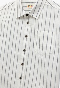 Camicia bianca a righe con strisce sottili blu scuro, dotata di una piccola tasca sul petto e bottoni marroni. Il tessuto è leggero e con texture.