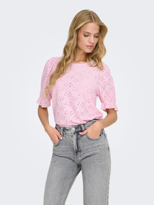 Ung kvinde med langt blondt hår iført en lyserød bluse med hulmønster og pufærmer samt højtaljede grå jeans, hænderne i lommerne, kigger ned.