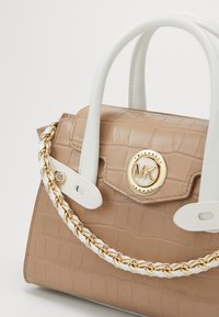 Vaaleanruskea krokotiilinnahkainen käsilaukku, jossa on valkoiset kahvat, kultaketjuakcentti ja brändin ympyrälogo etuosassa.