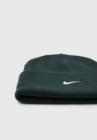 Tamna zelena pletena kapa s rebrastom teksturom, na prednjoj strani ima bijeli Nike swoosh logo. Preklopljena je na donjem rubu.
