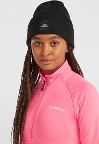 Chaqueta rosa con cremallera y un alto cuello, y gorro negro con un logo blanco. Pelo largo trenzado visible, fondo simple.