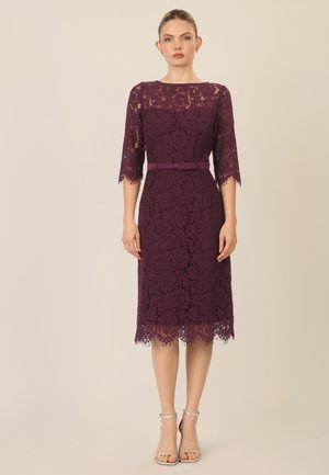 Spitzenkleid in tiefem Lila mit Dreiviertelärmeln, tailliertem Schnitt und gewelltem Saum. Begleitet von einem passenden Gürtel und silbernen Absätzen.