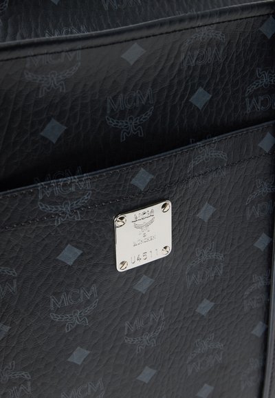 Μαύρο πορτοφόλι από δερμάτινη υφή με ανάγλυφο μοτίβο λογότυπου MCM, πλακέτα ασημένιου μετάλλου με χαραγμένα σημάδια και διαμαντένιες λεπτομέρειες.