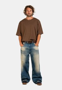 Man with curly hair stands with hands in pockets wearing oversized brown t-shirt, faded wide-leg jeans, brown belt, and dark shoes.