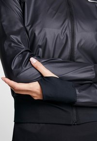 Veste de sport noire avec une texture lisse et légère, équipée d'une fermeture éclair et d'ouvertures pour les pouces sur les manches pour une fonctionnalité accrue.