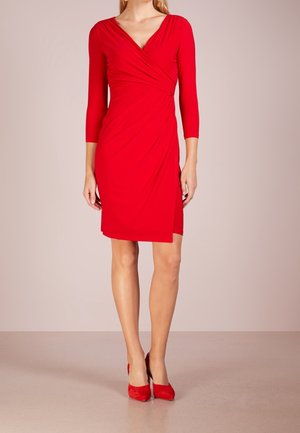 Kvinde iført en knælange rød slå-om-kjole med trekvartlange ærmer og røde højhælede sko, stående mod en ensfarvet baggrund.