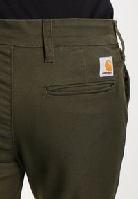 Olivgröna byxor av hållbart tyg, med en bakficka och ett rektangulärt Carhartt-logomärke i orange och vitt.