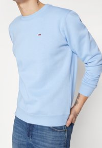 Personne portant un sweat-shirt bleu clair à col rond avec un petit logo sur la poitrine et un pantalon en jean bleu, avec une main tatouée reposant dans la poche arrière.