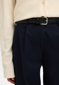 Άτομο που φοράει μια κρεμ μακρυμάνικη μπλούζα κλεισμένη μέσα σε ψηλόμεσο σκούρο μπλε παντελόνι με μαύρη δερμάτινη ζώνη.