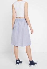 Bílý bezrukávový crop top s vlnitými okraji vypadá skvěle s modro-bílou pruhovanou sukní po kolena a navíc je doplněn námořnickými vysokými teniskami.