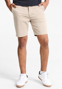 Beżowe bawełniane szorty o prostym kroju, z przednimi kieszeniami i gładką fakturą, zestawione z białymi sneakersami z czarnym akcentem.