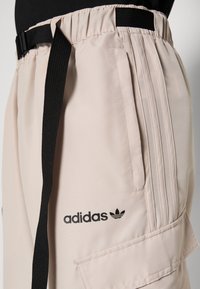 Svetle růžové kalhoty Adidas s černým páskem, s třemi pruhy, bočními kapsami a logem na stehně. Hladká tkanina s uvolněným střihem.