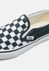 Chaussure Vans à enfiler avec un motif damier noir et blanc, semelle blanche et logo Vans sur une étiquette latérale.