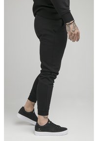 Černé tepláky s úzkým střihem, žebrovanými manžetami a předními kapsami. Kombinované s černými teniskami s bílými podrážkami a zlatými detaily.