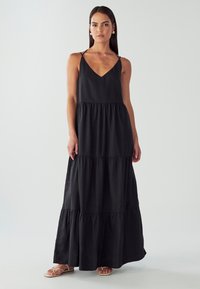 Žena stojící v černých dlouhých šatech s tenkými ramínky a volány, v béžových plochých sandálech na pozadí světlejednoduchého pozadí.