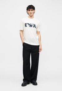 Άντρας με σγουρά μαλλιά στέκεται με τα χέρια στις τσέπες, φοράει ένα λευκό μπλουζάκι με σχέδιο, μαύρο παντελόνι με φαρδιά πόδια και μαύρα παπούτσια.